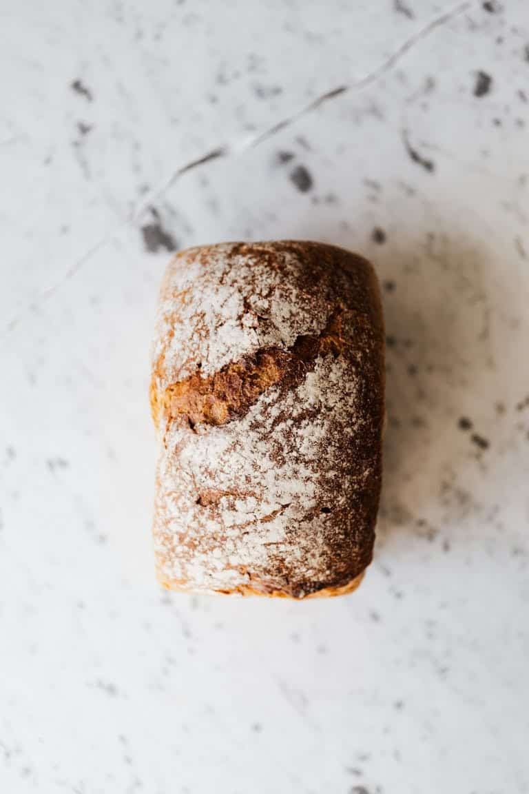 Top view of small rectangular baked bread loaf with uneven cracky surface and flour on top on marble table with spots